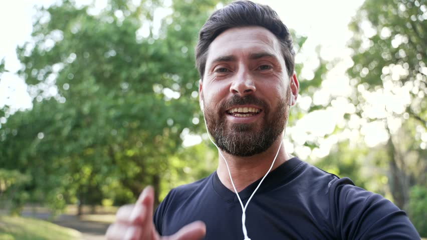POV Handsome sportsman communicates on a video call using a phone while walking in an urban city park. Smiling man with beard wearing earbuds waving, talking with a friend looking at the camera