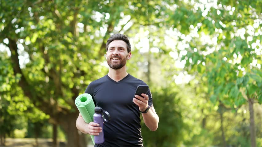 Sportsman engages in fitness by walking in park with yoga mat and water bottle, checking phone for workout updates. Smiling bearded athletic returning from training. Active lifestyle, healthy routine