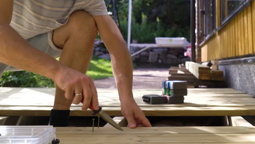 A man builds a wooden terrace in the backyard of a cozy house on a sunny summer day. He uses a drill and other tools while working on the decking. Surrounded by lush greenery and natural light.