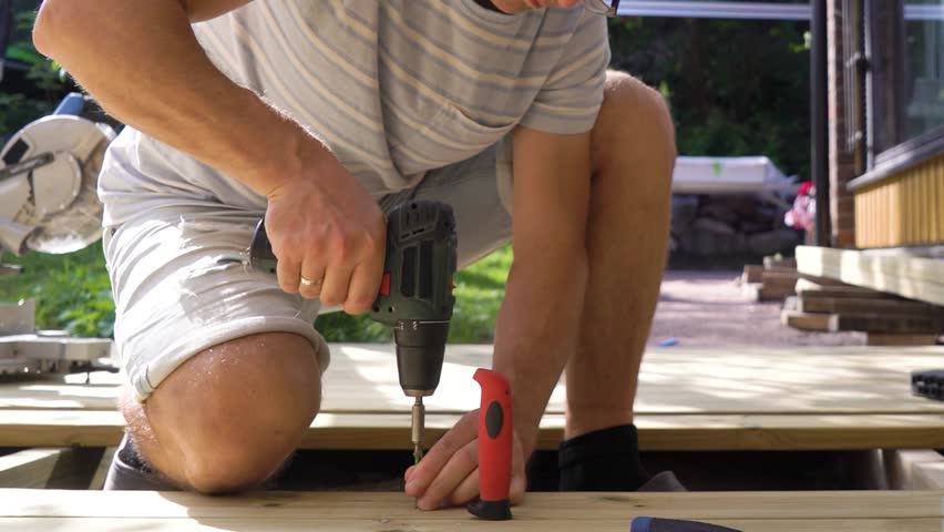 A man builds a wooden terrace in the backyard of a cozy house on a sunny summer day. He uses a drill and other tools while working on the decking. Surrounded by lush greenery and natural light.