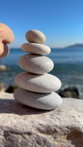 CLOSE UP, VERTICAL: Human hand knocks over a carefully balanced stack of white pebbles near the sea. Blue calm ocean contrasts with sudden action, symbolizing disruption and fragility of balance.