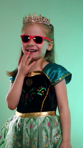 Vertical. A little princess poses for the camera, laughs and dances funny. Elegant young queen radiates joy and mischief. Cheerful movements of a girl in a crown and dress. Happy child