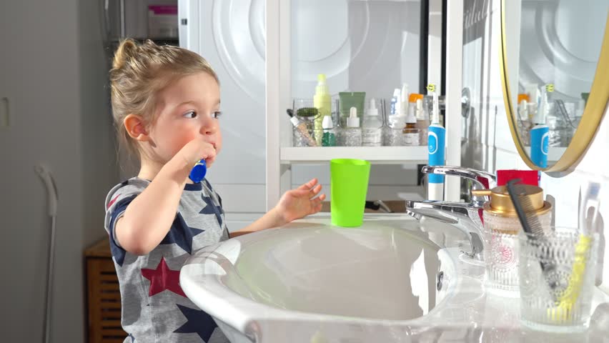 A toddler boy in a bathroom brushes his teeth while looking in the mirror. He holds a toothbrush and a green cup, learning good hygiene habits in a bright and modern home setting.