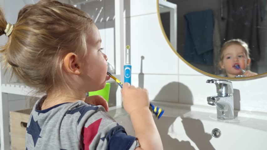 A toddler boy in a bathroom brushes his teeth while looking in the mirror. He holds a toothbrush and a green cup, learning good hygiene habits in a bright and modern home setting.