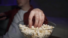 Comfortable man enjoying a movie night, using a remote control and grabbing popcorn from a glass bowl while sitting on a sofa in a dimly lit living room - Powered by Shutterstock - Get 15% off with code: PIKWIZARD15
