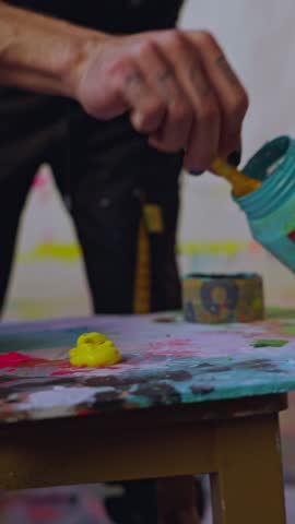 Close-up of artist applying blue paint to palette, vertical video. Hands of artist placing thick blue paint on messy palette in colorful studio environment.