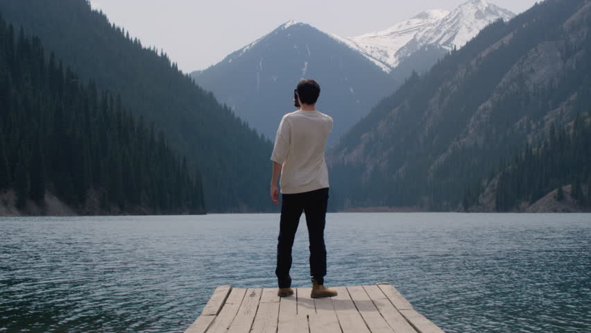 A man on a wooden pier on a mountain lake. A guy travels alone among the mountain peaks, an adventure. Male uses a smartphone, takes selfies. Blue water in the lake, untouched nature
