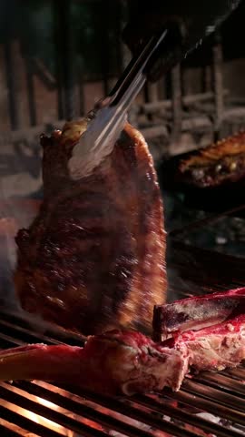 Juicy beef ribeye and bone in rib section sizzling on a hot grill, being expertly turned by a chef with tongs in a restaurant kitchen