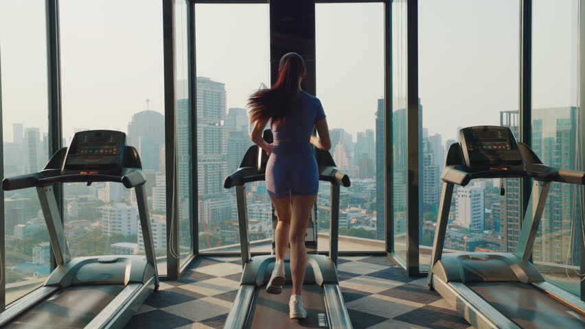 Athletic woman runs on treadmill near large windows with stunning city skyline. Sportive female exercises on trainer in luxury sports. Sunlight invigorates ambiance for workout