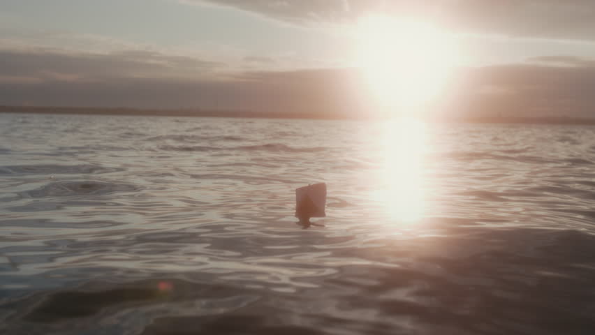 Full no people shot with lens flare of small white origami paper boat sailing and rocking on rolling lake waves in glaring sun during sunset