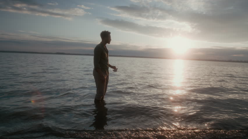 Wide shot with lens flare of young African American guy standing in lake water at sunset, launching origami paper boat, then looking at it sail away on rolling waves, in glaring sun