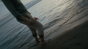 Medium rolling tracking shot of pensive young black man with bare feet walking on sandy lake beach, carrying bright orange daisy flower and smelling it, feeling lonely - Powered by Shutterstock - Get 15% off with code: PIKWIZARD15