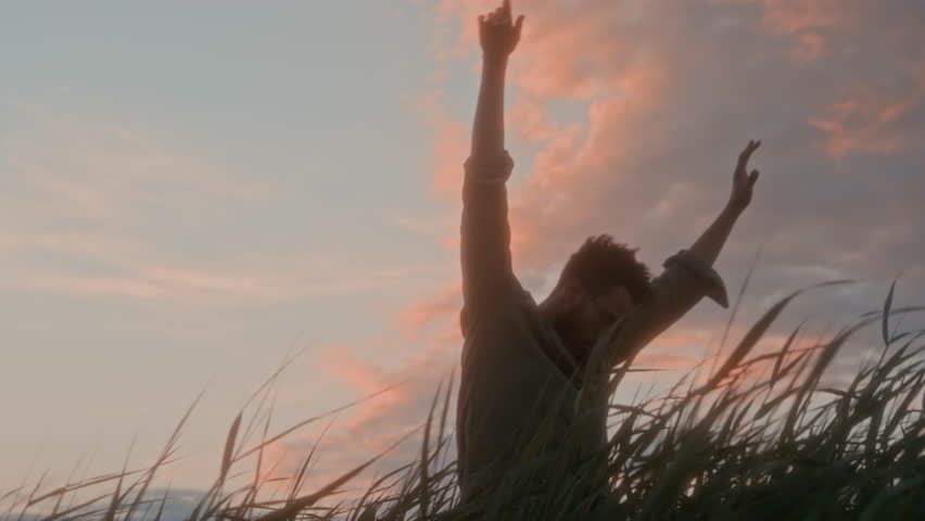 Medium low-angle shot of young man holding arms above head and swinging them in rhythm with wind, while performing meditative dance in green field after sunset, against pink clouds