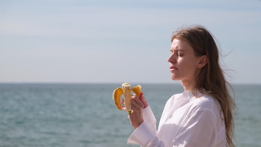 young woman walking on the beach in cool and windy weather and eating banana