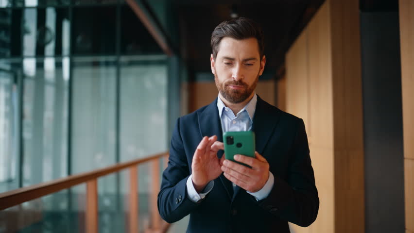 Smiling entrepreneur reading smartphone message walking hallway modern company closeup. Happy businessman in elegant suit messaging cellphone on go. Bearded manager receiving sms on mobile phone.