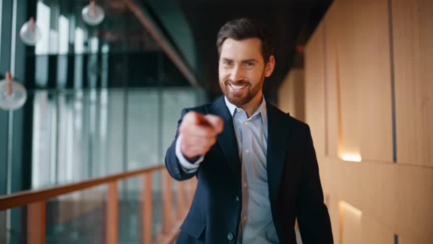 Successful businessman walking corridor modern company pointing finger to camera closeup. Confident bearded boss smiling buttoning elegant jacket. Luxurious happy executive going office hallway alone.