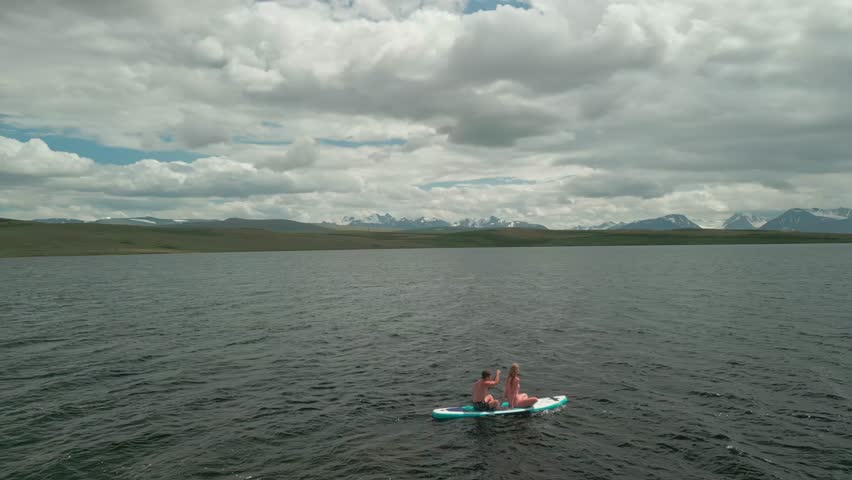 A young married couple swims on a SUP board in a mountain lake against a backdrop of green hills and mountains covered with snow. People are boating in a deserted lake in the mountains.