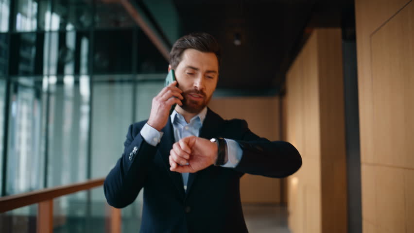 Happy business owner calling cellphone walking modern corridor closeup. Confident businessman negotiating by smartphone on go company hallway. Smiling boss talking mobile phone checking time on watch.