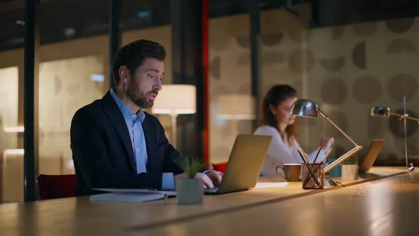 Bearded businessman talking to colleague working together at night coworking space closeup. Two coworkers focused on laptops sitting dark office. Smart employee explaining project to woman manager.