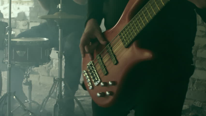 Muscular rock band members performing intense musical collaboration inside dilapidated industrial space, generating powerful rhythmic energy through bass guitar and drum interaction