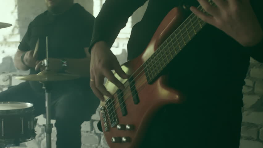 Muscular rock band members performing intense musical collaboration inside dilapidated industrial space, generating powerful rhythmic energy through bass guitar and drum interaction