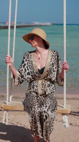 Pretty woman in sunglasses and straw hat having fun on beach swing. Smiling female tourist in leopard dress swings on the beach at summer vacation.