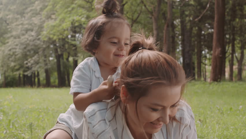 Medium shot of cute playful toddler boy having fun with mom lying on grass in park on warm sunny day