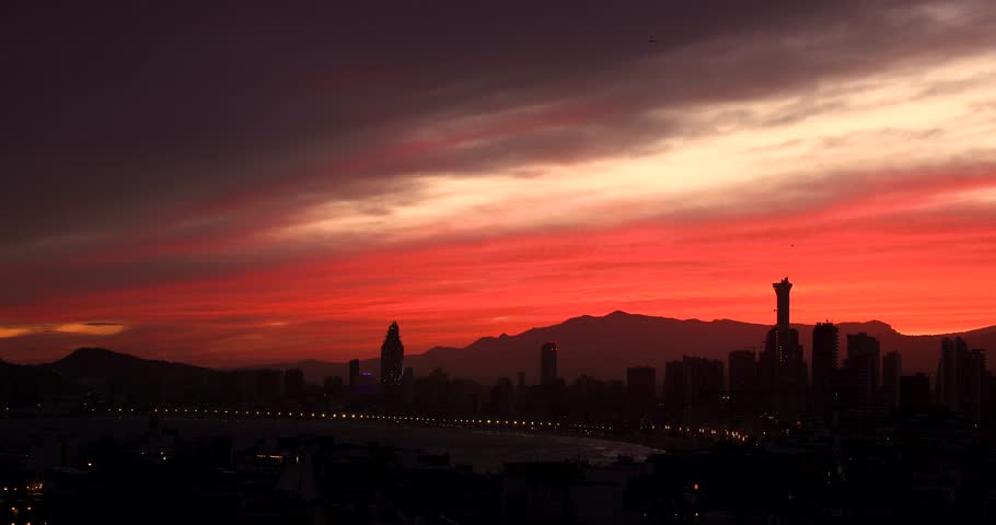 Footage of the sun setting in the evening over the old town of Benidorm in Spain showing the beautiful red sun set with clouds in the sky along side high rise apartments in the summer time