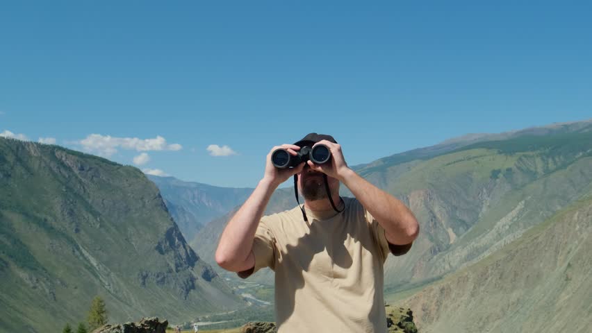 A man traveler looking through binoculars standing on top of mountain with blue sky on the background.