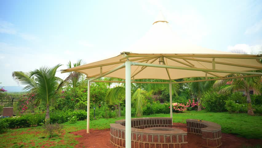 Circular Stone bench sits under canopy in tropical garden, Hexagonal gazebo stands at center of park, Shaded patio surround by palms and lush plants, Leafy trees around open lawn space, Red soil