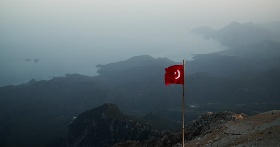 The Turkish flag flies high on a flagpole on Mount Tahtali in sunset light with a misty sea coast in the background. Wide aerial view.