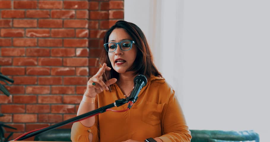 Indian Asian young entrepreneur confidently delivering speech to aspiring business professionals at conference, standing at the podium on mic as her team joins from behind, clapping and cheering