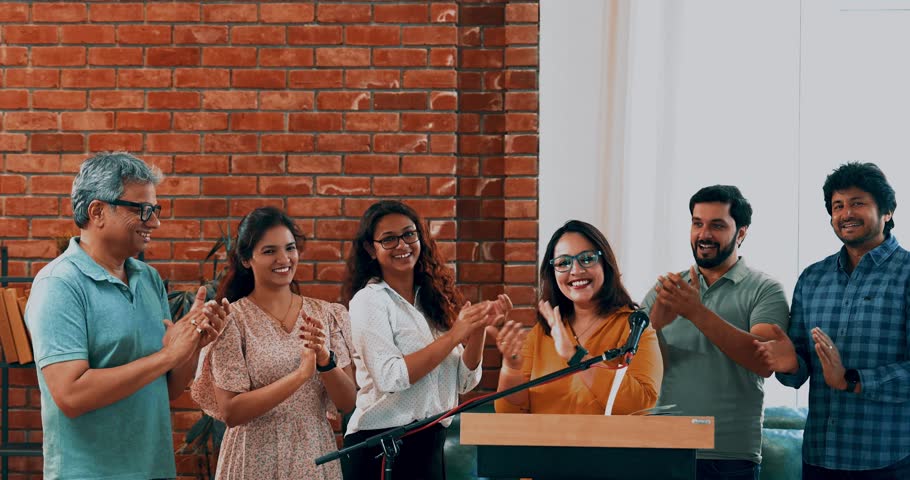 Indian Asian young entrepreneur confidently delivering speech to aspiring business professionals at conference, standing at the podium on mic as her team joins from behind, clapping and cheering