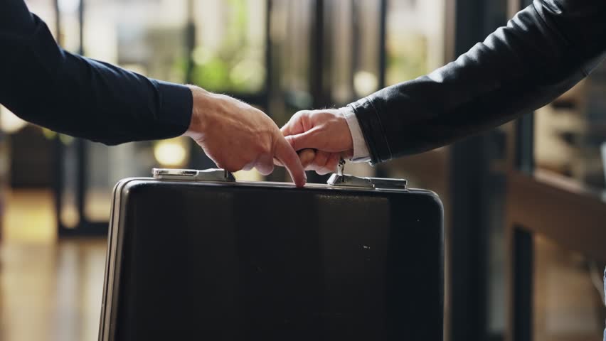 Suited man hands over a suitcase to another, secrecy and suspicion. Government conspiracy or corporate fraud and political corruption.Handshake finalizes transaction. Power and deception collusion