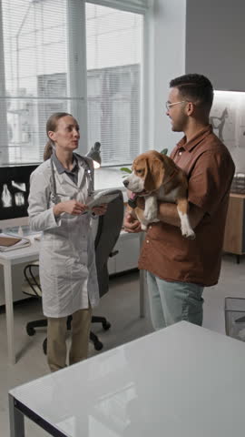 Vertical shot of female veterinarian holding medical records and consulting male pet owner holding beagle dog in examination room of vet clinic