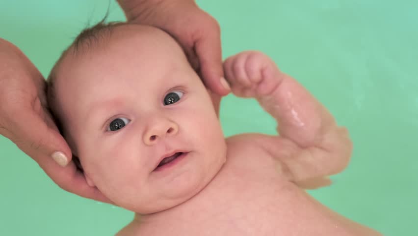  SLOW MOTION: Mom teaches a newborn baby to swim and stay on the water. Two-month-old baby is bathing in a large bathtub.   Newborn baby is in the water, bathroom. Hygiene procedures of a newborn boy