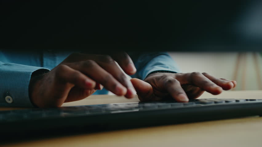 Focused hands of African American man wearing blue shirt typing on keyboard while working at desk. Rapidly inputting data or composing message in professional setting with modern workspace elements.