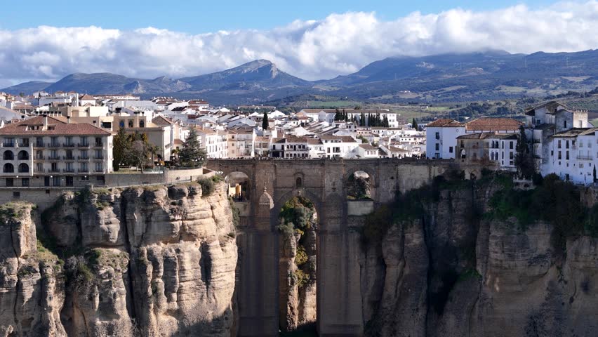 Ronda, Spain, a breathtaking panning aerial view capturing the iconic Puente Nuevo bridge, dramatic cliffs, charming white houses, and a picturesque cloudy sky creating a captivating landscape