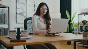 Elegant Caucasian woman with long brown hair and glasses sitting at desk and turning head while smiling slightly. Resting hands on laptop keyboard while pausing from work. Bright modern office. - Powered by Shutterstock - Get 15% off with code: PIKWIZARD15