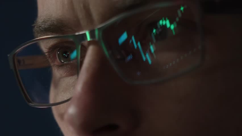 Close-up of a focused trader wearing glasses with a glowing stock market chart reflection. Serious crypto investor analyzing the cryptocurrency market. Finance, trading, and investment concept.