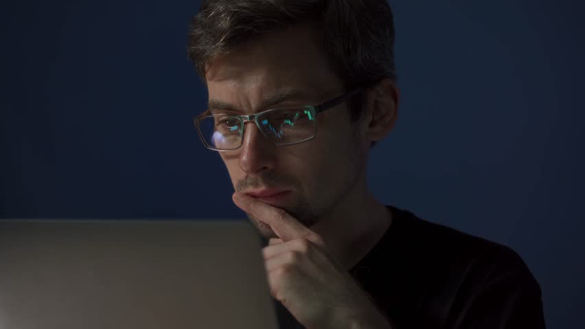 A smiling crypto trader wearing glasses clenches fists in excitement while looking at stock market charts reflected in his lenses on a laptop screen. Happy trader celebrating stock market success 