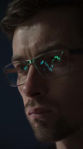 Close-up of a focused trader wearing glasses with a glowing stock market chart reflection. Serious crypto investor analyzing the cryptocurrency market. Finance, trading, and investment. Vertical video