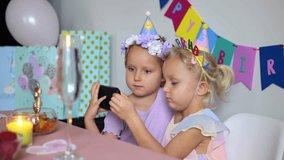 Two young sisters wearing colorful party hats sitting at decorated table, sharing smartphone screen during birthday celebration, enjoying digital connection and moment of togetherness - Powered by Shutterstock - Get 15% off with code: PIKWIZARD15