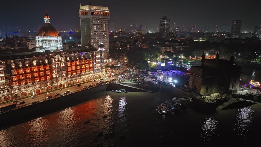 Aerial night view of the Gateway of India. Cityscape of old Mumbai town. Evening weather on the seashore of Mumbai. Cinematic view of the Taj Palace Hotel. Fishing boats sail in the sea in the evening