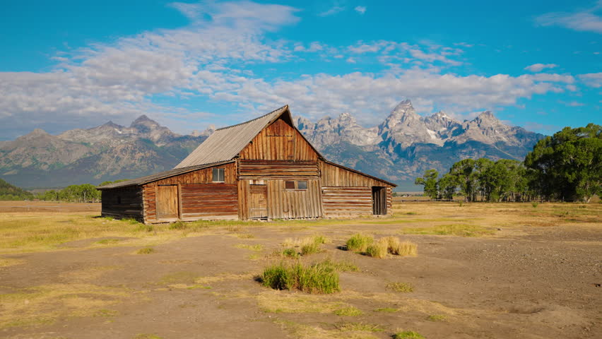 Sweeping panning shot captures a historic wooden barn with a breathtaking mountain range backdrop, showcasing rustic charm and scenic beauty.