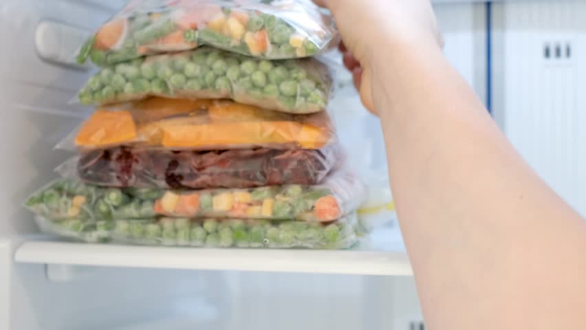 Female hand holding zip bag with frozen green peas and carrots against open freezer background.