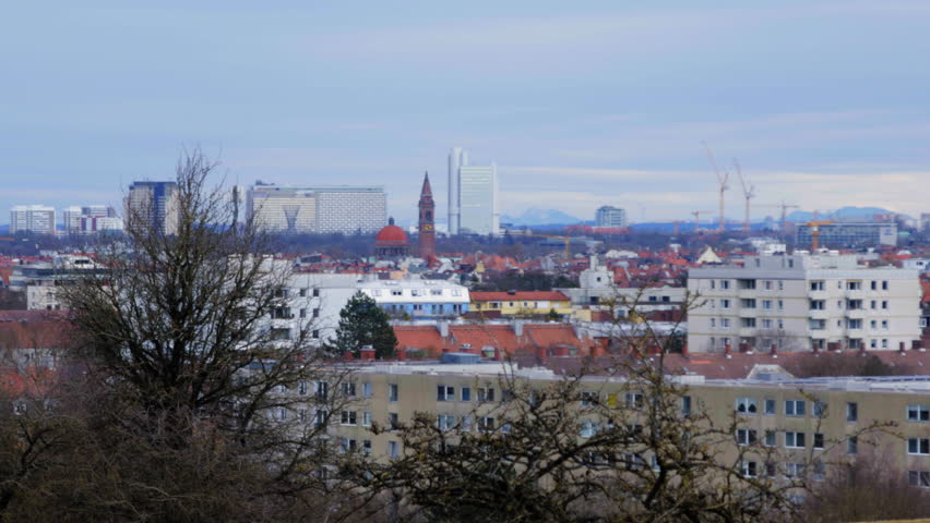 A panoramic view of Munich, Germany, showcasing a mix of historic and modern architecture with the Alps in the background. Construction cranes indicate urban development under a cloudy winter sky.