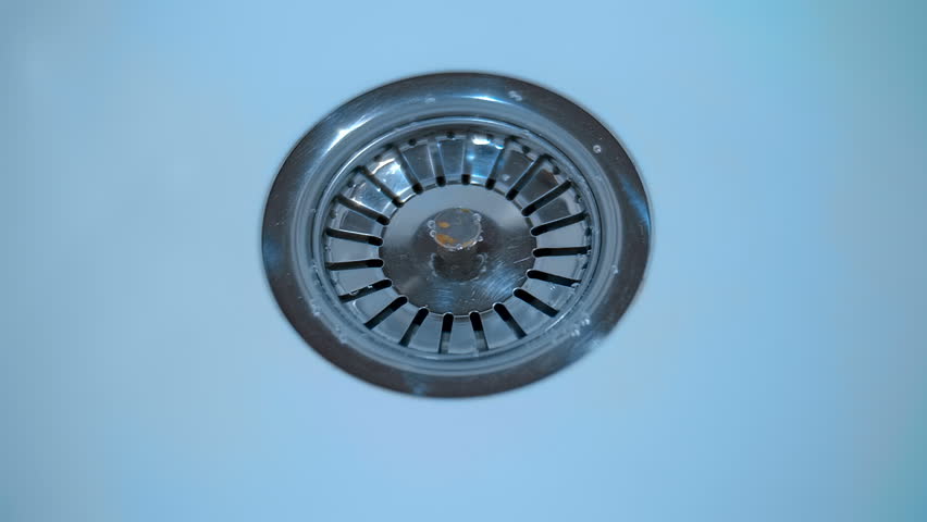 A stream of water flows into a shiny chrome drain in a white sink. Closeup. Macro. Slow mo, slow motion, high speed camera, 250fps