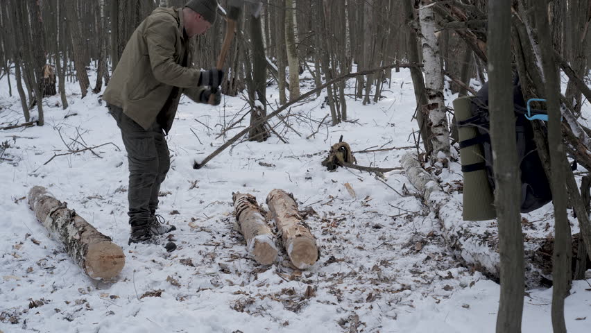 A man in winter clothing chops firewood in a snowy forest with an axe. His backpack and outdoor gear suggest bushcraft, survival, and off-grid living.