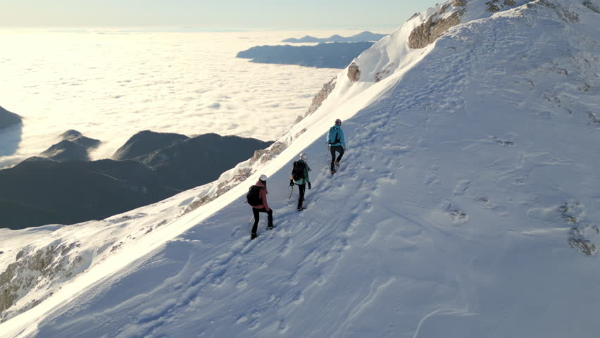 Three climbers ascend a snowy mountain ridge, their joyous journey highlighted by the breathtaking view of clouds and distant peaks.